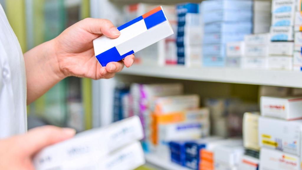 A hand holding a medicine packet with pharmacy shelves in the background