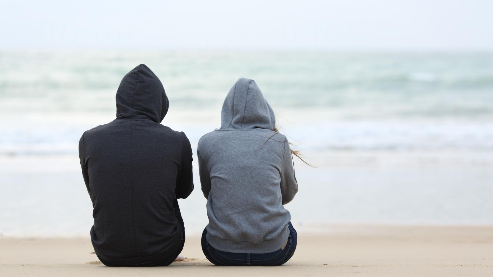 a view from behind of two people sitting on a beach looking our to sea. Both are wearing hooded sweatshirts