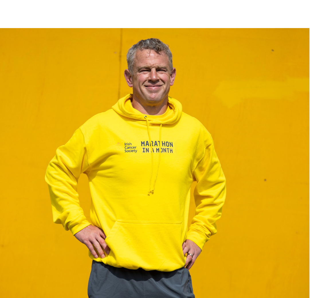 A man stands against a yellow background, in a slightly darker yellow hoodie with "Marathon in a Month" printed across the chest