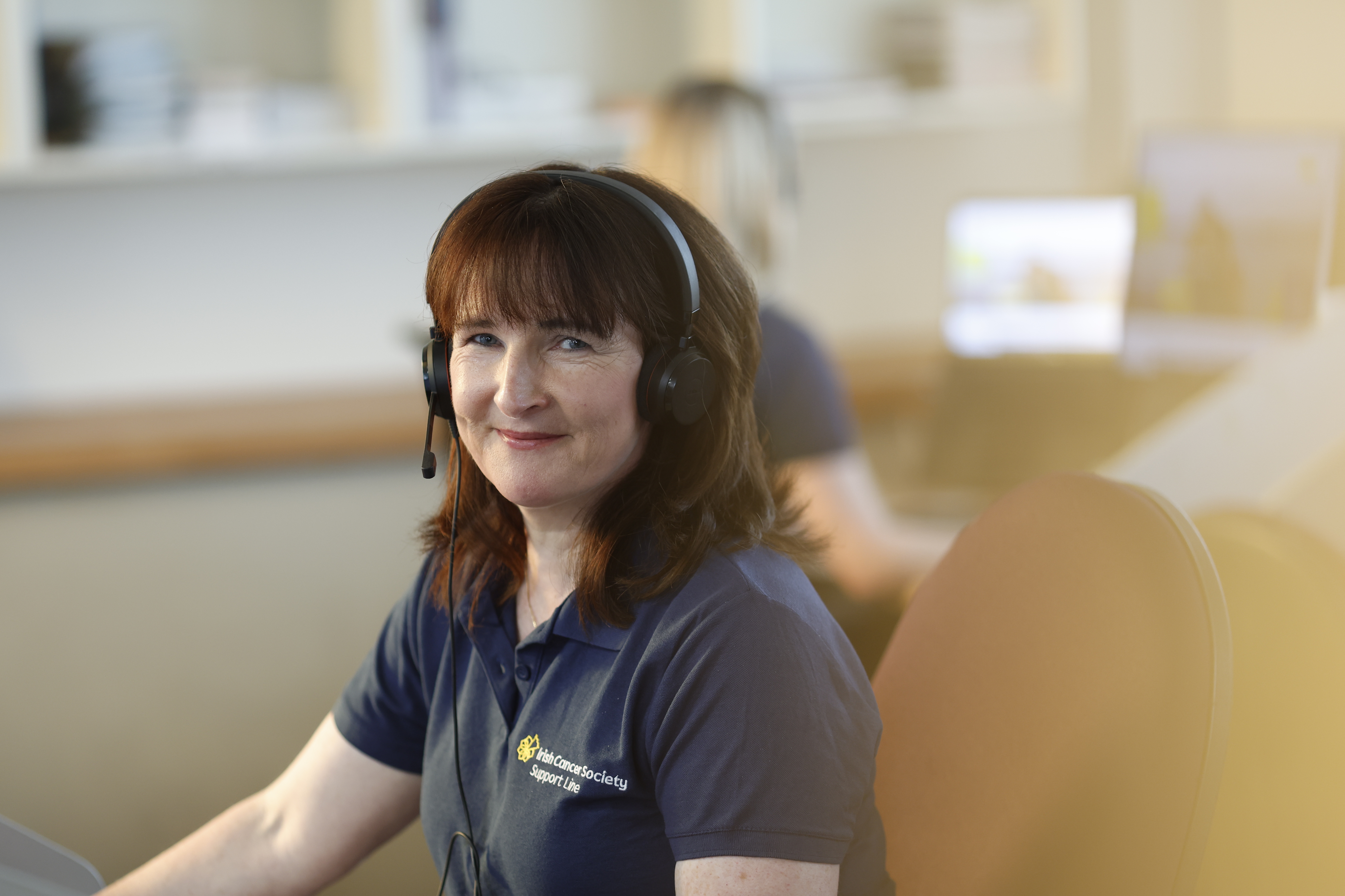Edel Brangan, Support Line Cancer Nurse. Edel looks direct to camera with a warm smile. Edel is wearing a headphone set, ready to receive calls.