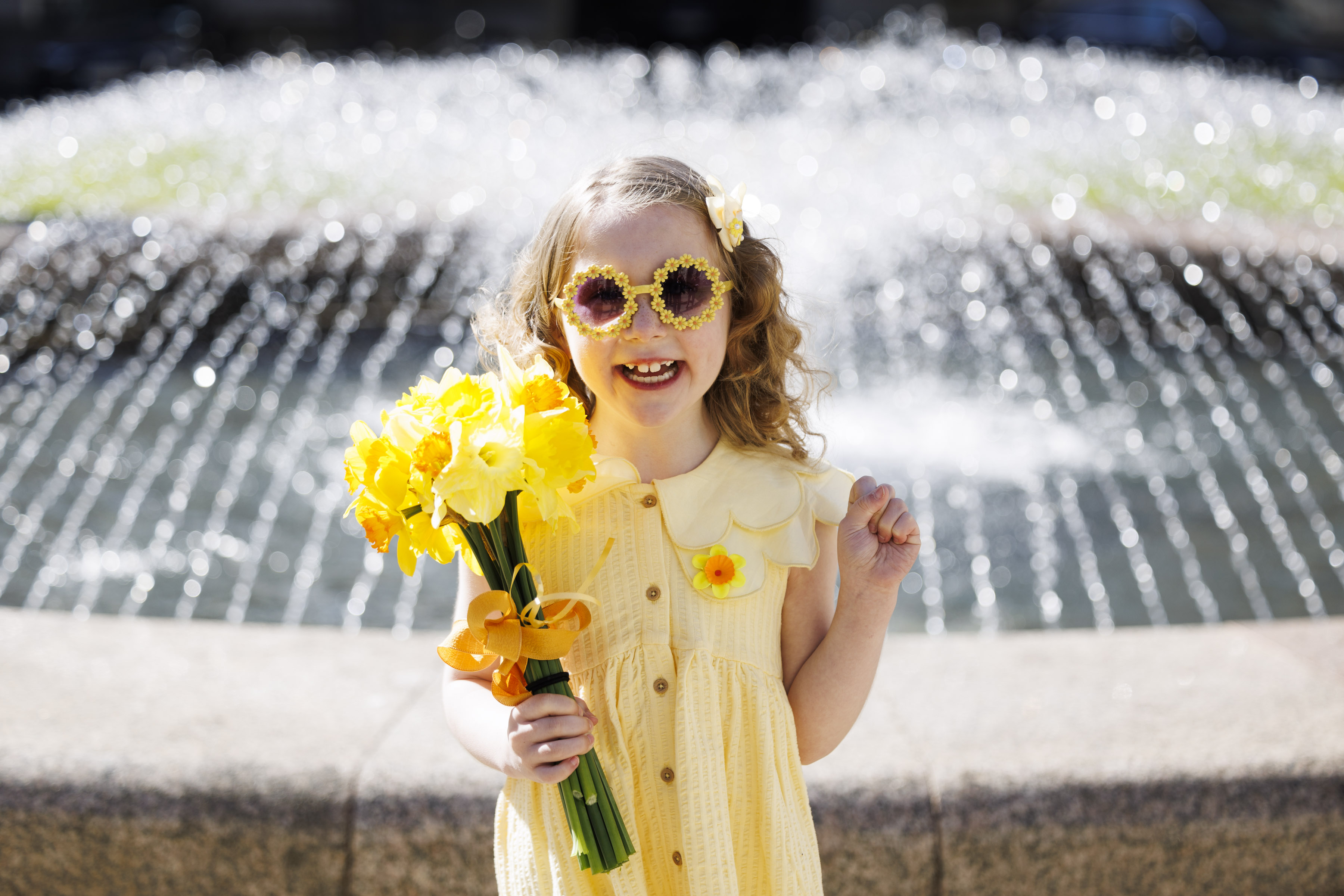 Daffodil Day image - little girl with sunglasses and in a yellow dress standing in front of the fountain
