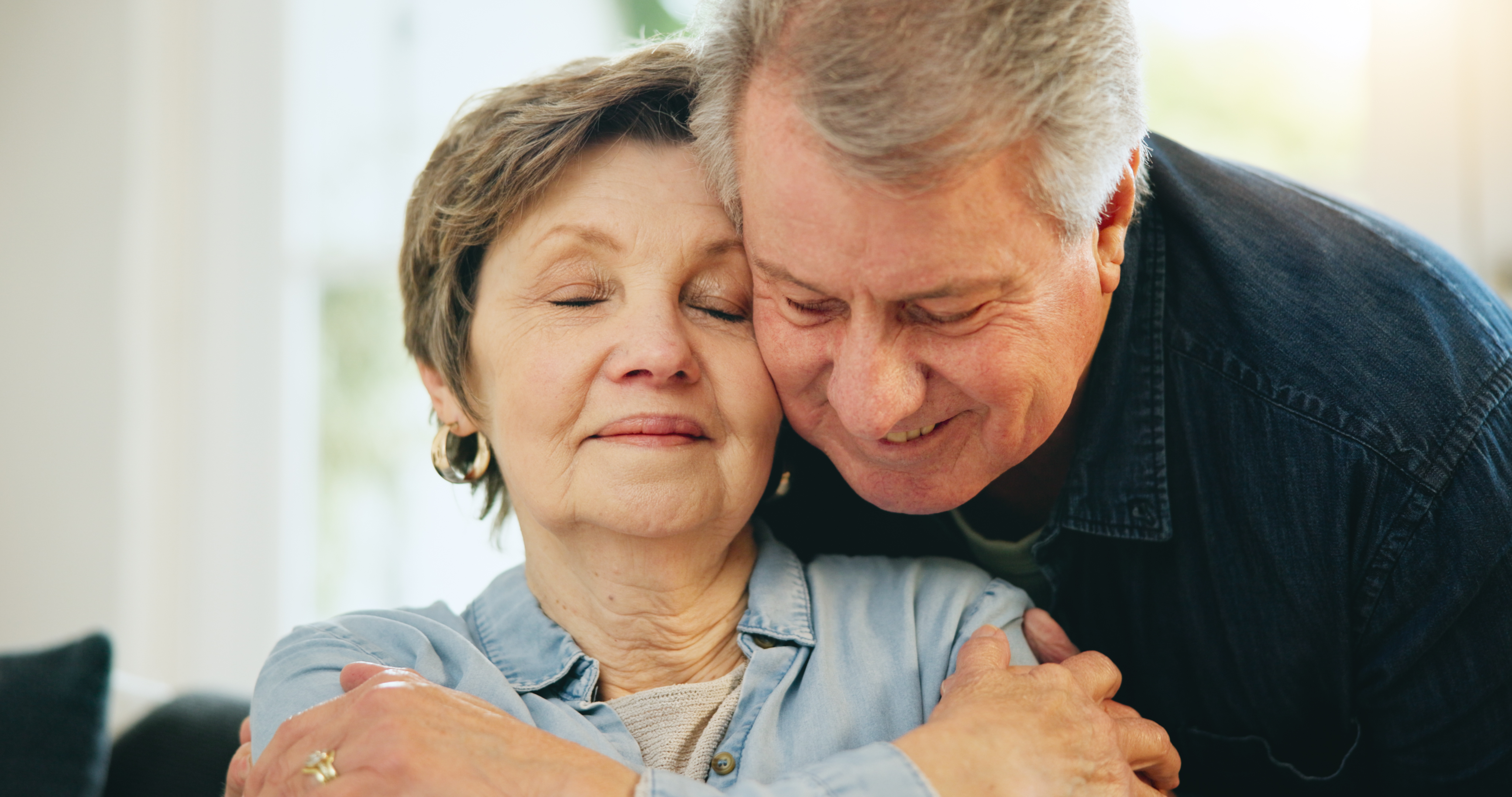 A mature man and woman hugging. The woman is sitting and the man has his arms around her shoulders and his face next to hers. She looks content. rriage partner. Romance, love and sweet elderly man, senior woman or old people hugging 
