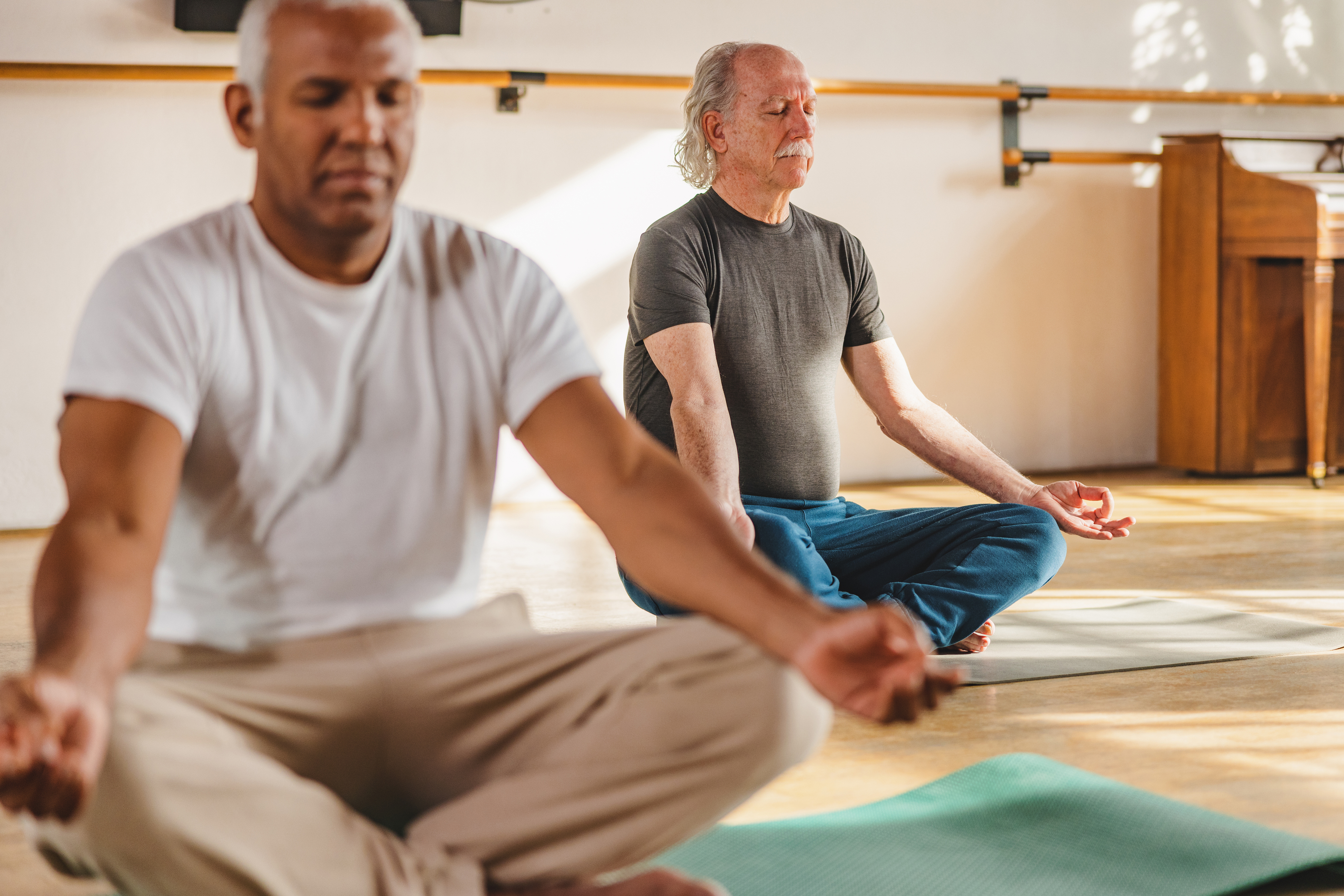 Three-quarter side shot with blurred background of two multicultural mature men yoga participants seated with crossed legs in their mats while meditating with closed eyes and open hands during a sunny day indoors.