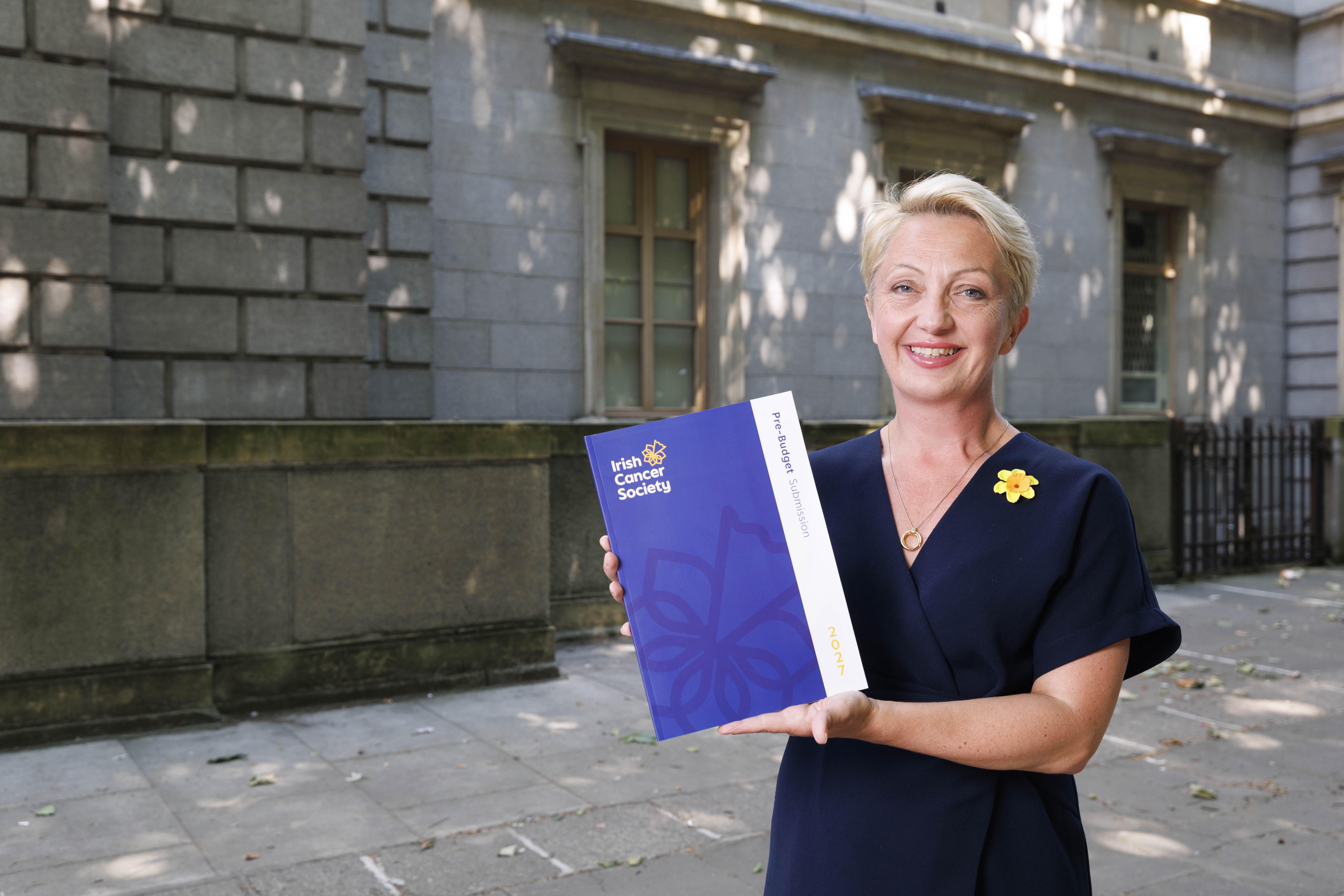Woman with short blond hair and a black dress on holding a booklet up to the camera with the title 'Pre-budget submission 2026'