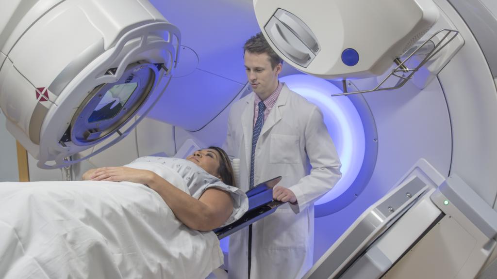 A lady is lying under a radiotherapy machine. A doctor is standing behind her head