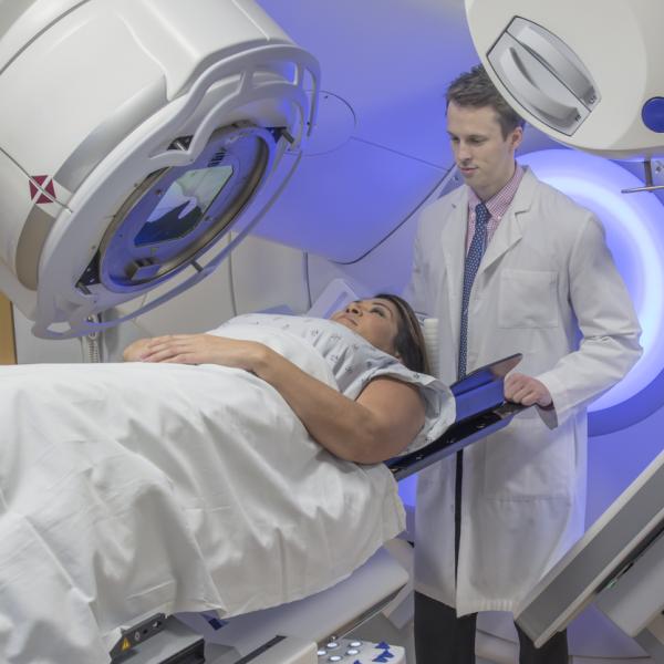 A lady is lying under a radiotherapy machine. A doctor is standing behind her head