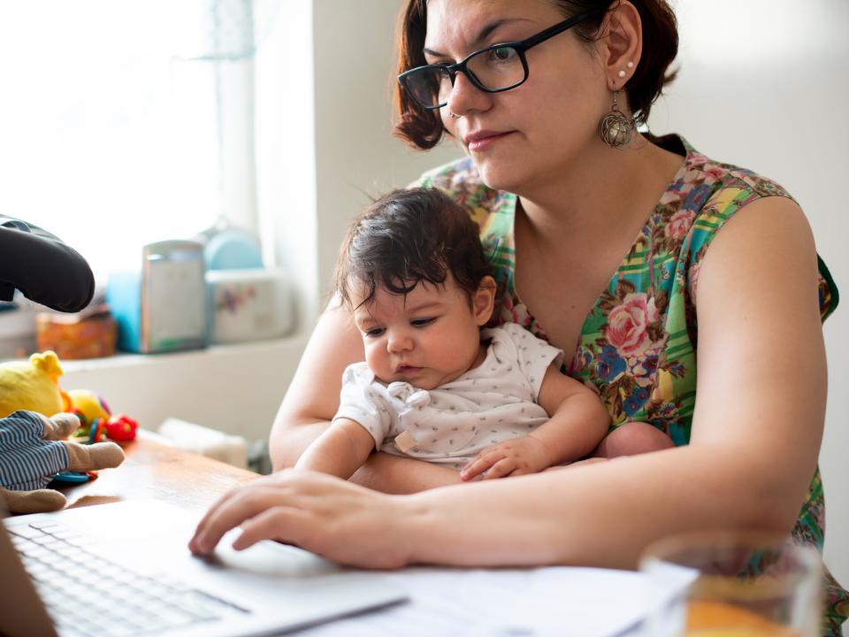 A mother uses her laptop with a baby on her lap