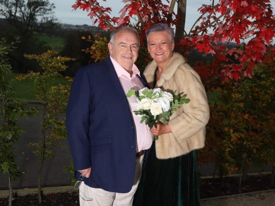 Maggie Donnelly pictured with her husband Pat in front of a tree with red leaves. Maggie is holding a bouquet of white flowers. 