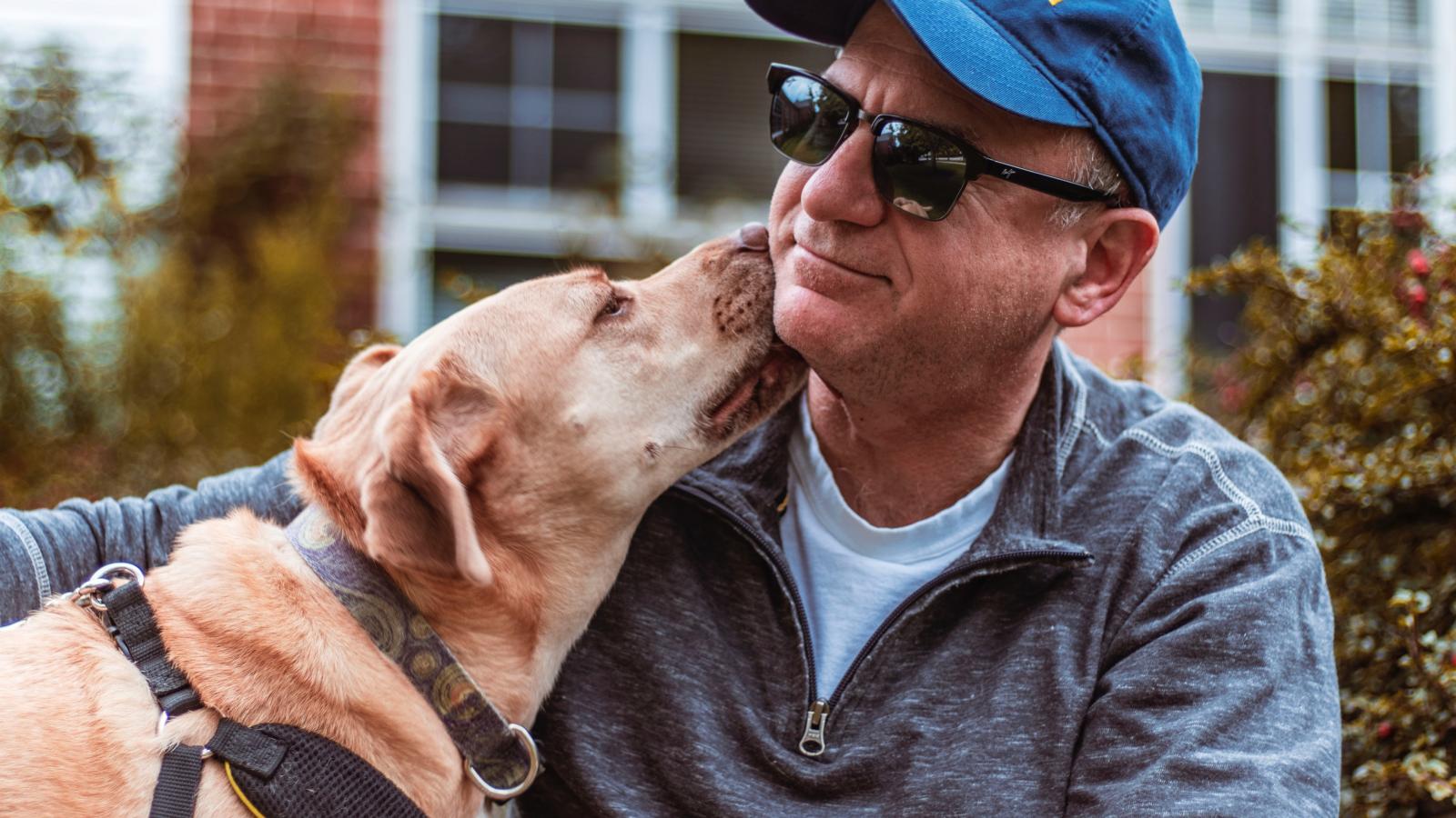 A man wearing dark glasses is being licked on the face by his dog