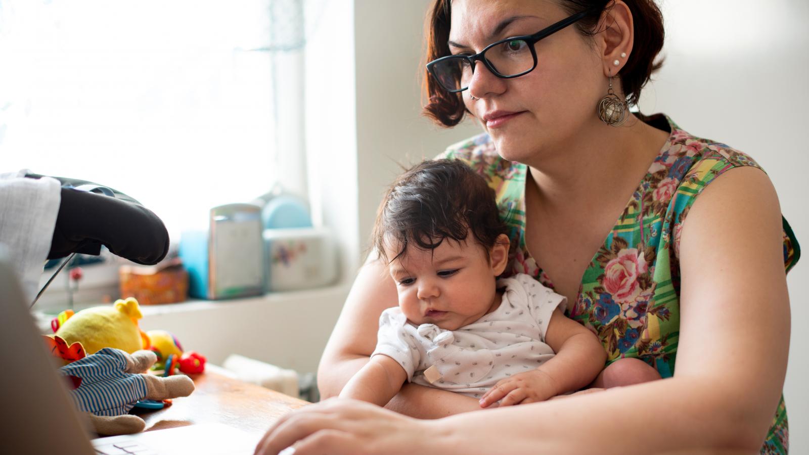 A mother uses her laptop with a baby on her lap