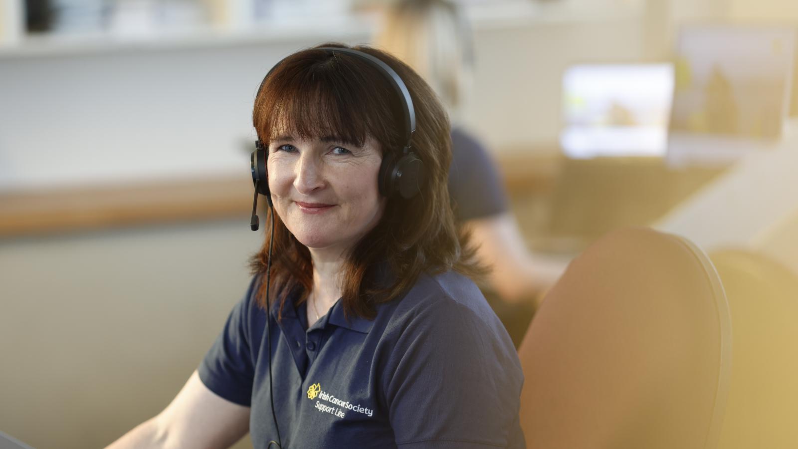 Edel Brangan, Support Line Cancer Nurse. Edel looks direct to camera with a warm smile. Edel is wearing a headphone set, ready to receive calls.