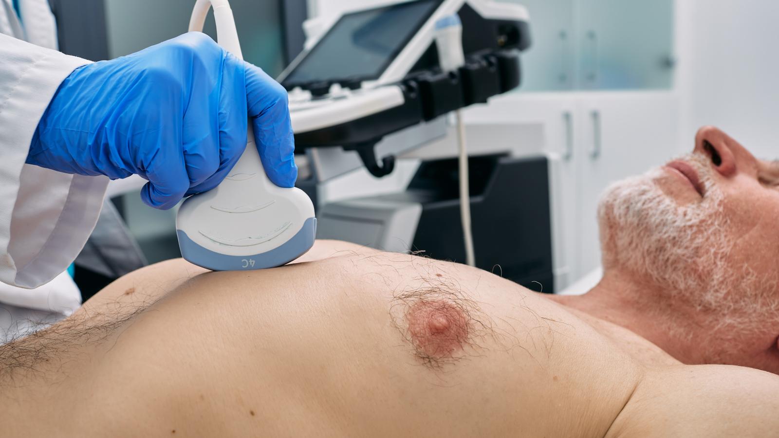 A middle aged man is bare chested on a examination bed. A female nurse is sitting beside him holding an ultrasound probe on his chest. 