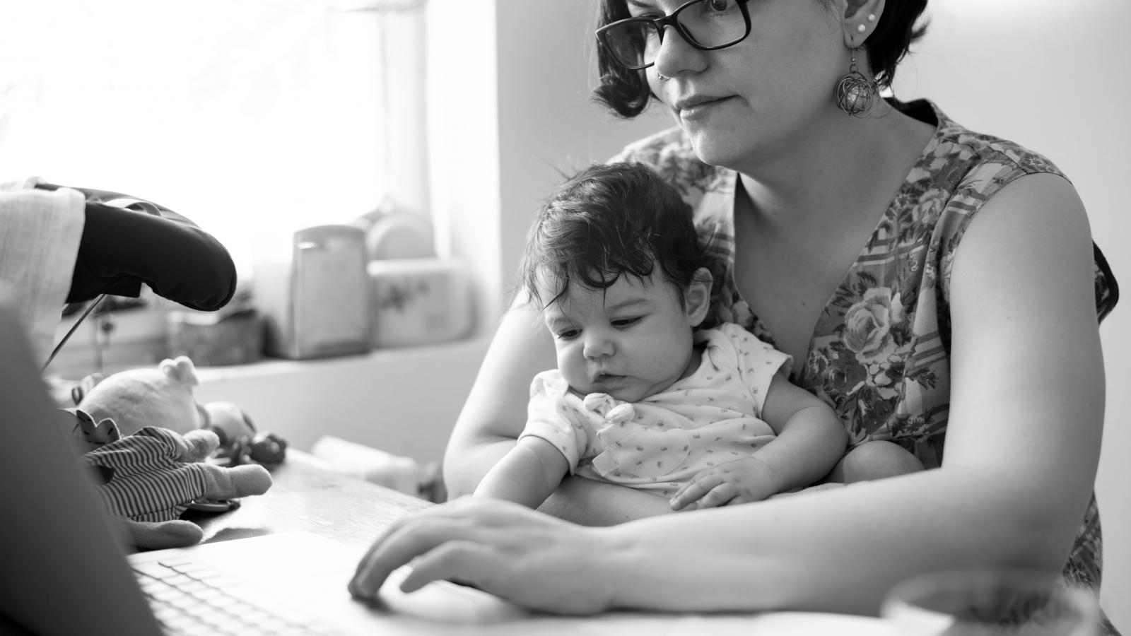 A mother uses her laptop with a baby on her lap